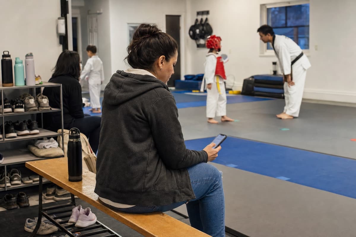 A parent checking a phone from the edge of a youth martial arts class